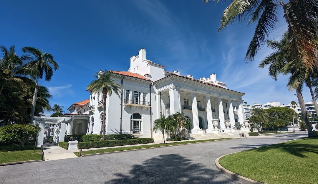 Exterior view of a luxury historic mansion with grand white columns, a tiled roof, and surrounding palm trees