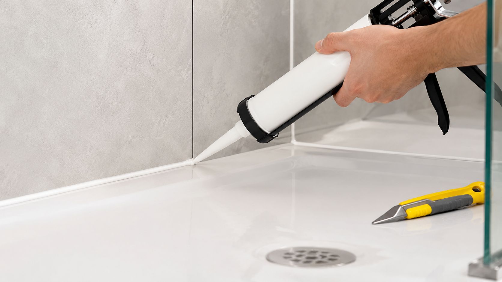 A hand using a caulk gun to apply white silicone sealant along a shower base edge