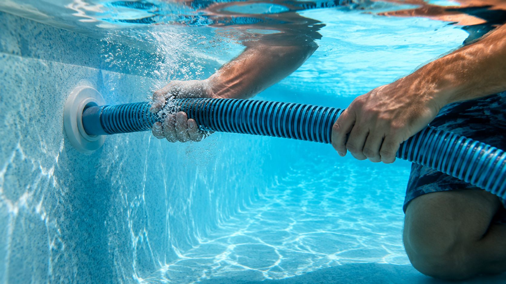 A person underwater connecting a blue pool vacuum hose to the filtration system port in a pool.